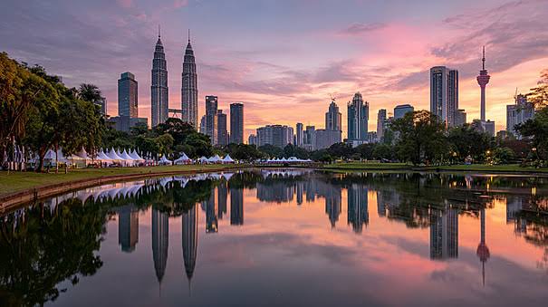 The Kuala Lumpur skyline at sunset, with the Petronas Towers and the KL Tower reflected in the still water of a lakeside park.
