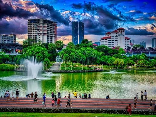 A fountain and tropical palms in a lakeside park in Kuala Lumpur, with city buildings and heavy cloud behind and visitors walking along the waterfront promenade.